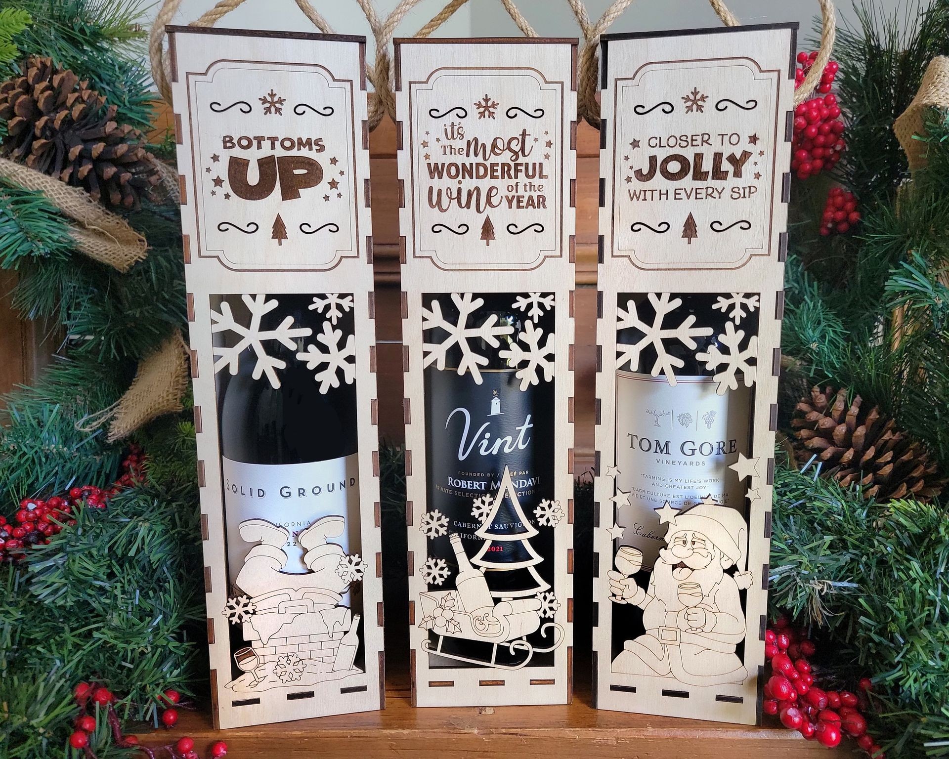 Three festive wine gift boxes with holiday designs and greenery in the background.