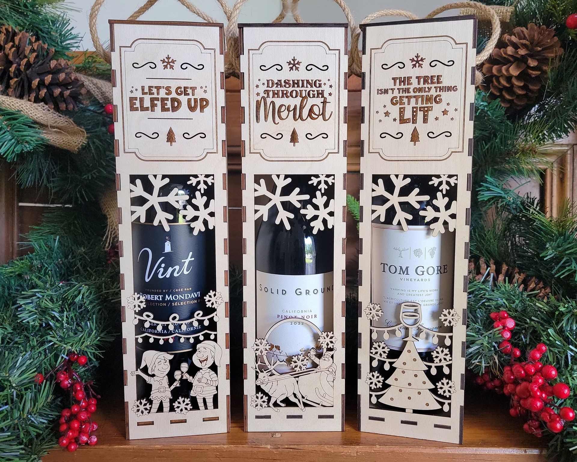 Three festive wine bottle gift boxes with holiday-themed designs and text, surrounded by pine and berries.